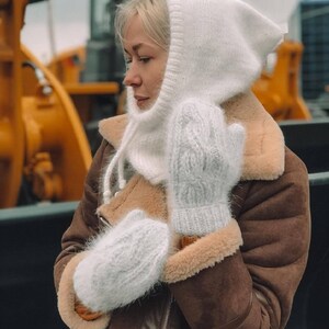 Pu&ograve; includere: Una sciarpa con cappuccio in maglia bianca e guanti abbinati. La sciarpa ha un design lungo e fluido e i guanti hanno un motivo a treccia.
