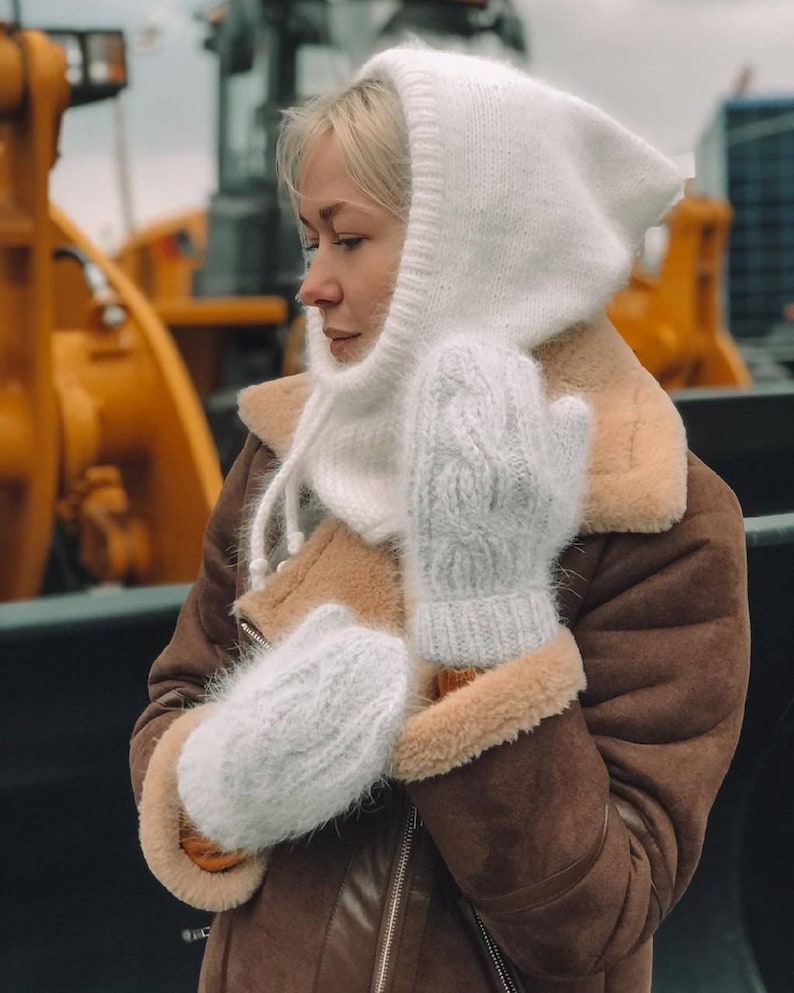 Pu&ograve; includere: Una sciarpa con cappuccio in maglia bianca e guanti abbinati. La sciarpa ha un design lungo e fluido e i guanti hanno un motivo a treccia.