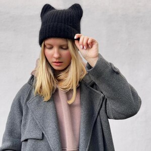 May include: A woman wearing a black cat ear beanie and a gray coat over a pink hoodie.