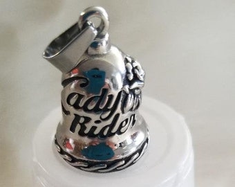 May include: A silver-colored bell pendant with the words "Lady Rider" engraved on the front. The bell is on a white surface with the number "1" in black below it. The pendant has a loop for a chain.