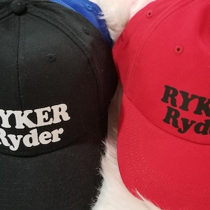 May include: Two baseball caps, one black with white text and one red with black text. Both caps have the name "Ryker Ryder" embroidered on the front.