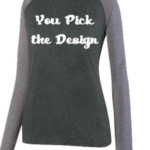 May include: A long-sleeved raglan shirt. The body is dark grey, with light grey sleeves. The text "You Pick the Design" is printed in white across the front. The shirt is made of a soft, comfortable material.