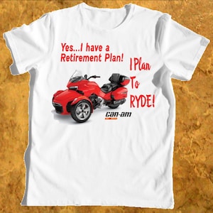 Może przedstawiać: Biała koszulka z nadrukiem czerwonego, czarnego i srebrnego trójkołowego motocykla z tekstem "Yes...I have a Retirement Plan! Plan To RYDE!" i "can-am" na niej.