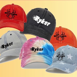 May include: Five different baseball caps with various designs. One red cap with a black spider logo, one black cap with a white flame and the word "Ryker", one orange cap with a black spider logo, one tie-dye cap with a white flame and the word "Ryker", and one blue denim cap with white stars and a black spider logo.