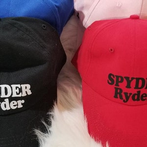 May include: A collection of baseball caps in various colors, including black, red, blue, and pink. Each cap features the text "SPYDER Ryder" in white or black lettering. The caps are displayed on a white, fluffy surface.
