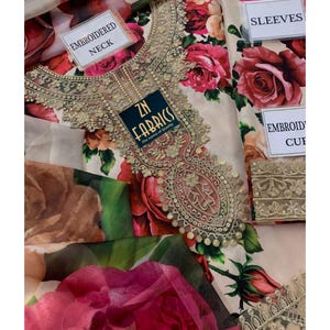 Puede incluir: Una tela floral crema con grandes rosas rojas. La tela presenta un escote bordado con detalles dorados. La etiqueta dice "ZN Fabrics". Etiquetas adicionales indican "Embroidered Neck", "Sleeves" y "Embroidered Cuff".