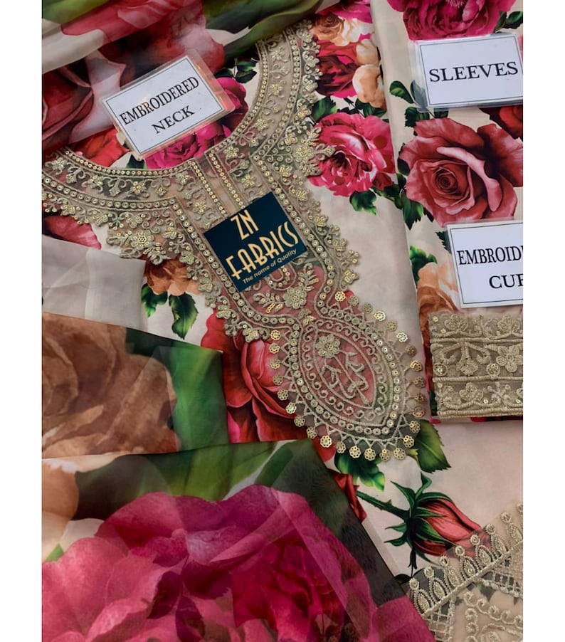 Puede incluir: Una tela floral crema con grandes rosas rojas. La tela presenta un escote bordado con detalles dorados. La etiqueta dice "ZN Fabrics". Etiquetas adicionales indican "Embroidered Neck", "Sleeves" y "Embroidered Cuff".