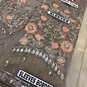 以下が含まれることがあります： ピーチとシルバーの色合いで複雑な花の刺繍が施されたグレーの生地のクローズアップ。 デザインにはスパンコールとパールのアクセントが含まれており、「SLEEVES」、「SLEEVES BORDER」、および「HAND WORK」と書かれたラベルが付いています。