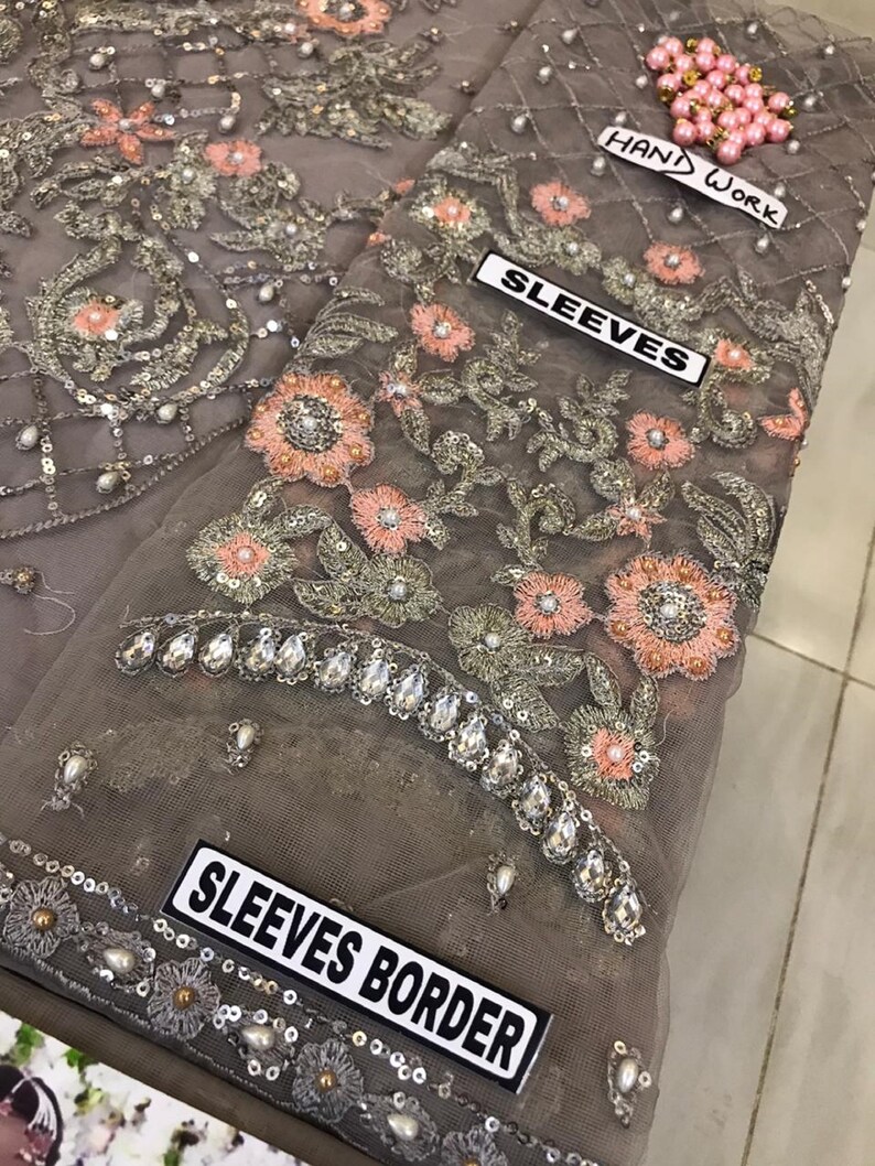 以下が含まれることがあります： ピーチとシルバーの色合いで複雑な花の刺繍が施されたグレーの生地のクローズアップ。 デザインにはスパンコールとパールのアクセントが含まれており、「SLEEVES」、「SLEEVES BORDER」、および「HAND WORK」と書かれたラベルが付いています。