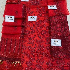 May include: A vibrant red embroidered fabric set, including a dupatta, trousers, and front and back panels. The fabric is adorned with intricate floral patterns and sequins. The set also includes embroidered sleeves. The labels read "FRONT", "BACK", "SLEEVES EMBROIDERED", "TROUSER", and "DUPATTA".