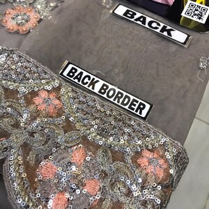 以下が含まれることがあります： グレーの生地のクローズアップ。複雑な刺繍とスパンコールのディテールが施されています。デザインは、ピーチ、シルバー、ブラウンの色合いのフローラルパターンが特徴です。「BACK BORDER」の文字が白いラベルに表示されています。