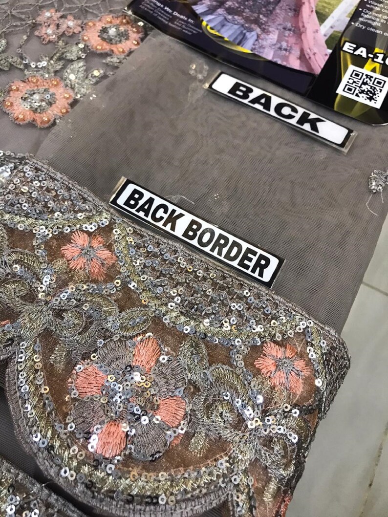 以下が含まれることがあります： グレーの生地のクローズアップ。複雑な刺繍とスパンコールのディテールが施されています。デザインは、ピーチ、シルバー、ブラウンの色合いのフローラルパターンが特徴です。「BACK BORDER」の文字が白いラベルに表示されています。
