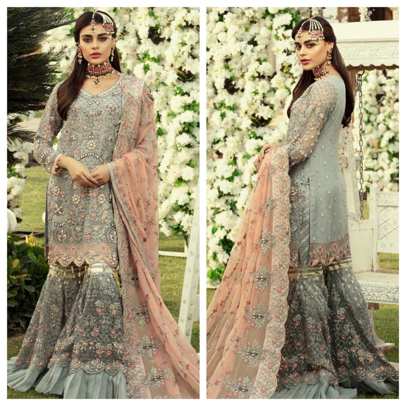 以下が含まれることがあります： グレーの刺繍入りフォーマルドレスと、お揃いのドゥパッタ。ドレスは複雑な花柄、ビーズ細工、フレアボトムが特徴です。ドゥパッタはソフトピーチ色で、繊細な刺繍が施されています。白い花の背景の前で屋外で着用されています。