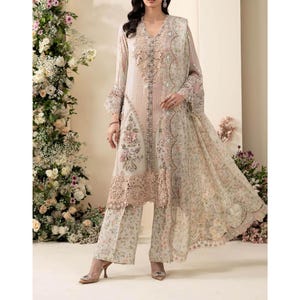 May include: A light beige long-sleeved tunic featuring intricate floral embroidery, sequin embellishments, and a scalloped lace hem. The outfit includes matching wide-leg trousers with a delicate floral print and a sheer, embroidered dupatta shawl.