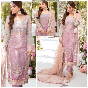 May include: A three-piece formal outfit in shades of pink and gray. The sheer tunic features intricate embroidery and long sleeves. The ensemble includes matching pants and a flowing scarf. The outfit is accessorized with a small, beaded clutch and high heels.