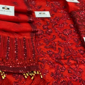 May include: Close-up of a vibrant red fabric with intricate floral embroidery and sequin embellishments. The fabric appears to be a sheer material, possibly a dupatta or other garment component. Tassels with gold accents add a decorative touch.