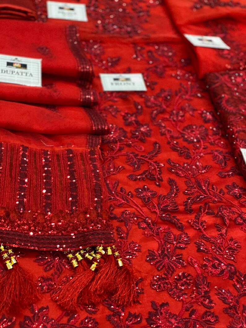 May include: Close-up of a vibrant red fabric with intricate floral embroidery and sequin embellishments. The fabric appears to be a sheer material, possibly a dupatta or other garment component. Tassels with gold accents add a decorative touch.