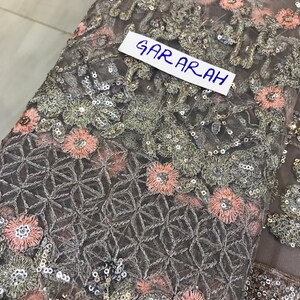 以下が含まれることがあります： 複雑な花の刺繍とスパンコール装飾が施されたグレーの生地のクローズアップ。ピンクとシルバーの花、幾何学模様、白いラベルに「GARARAH」の文字がデザインされています。生地は繊細で透け感があります。