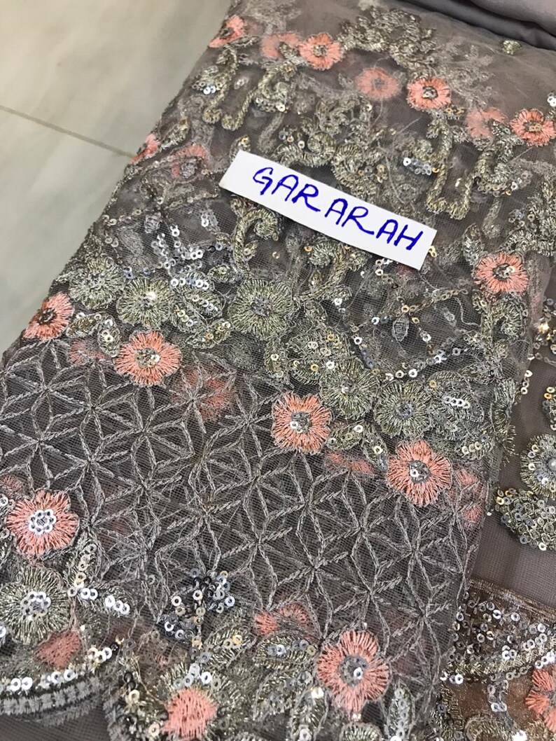 以下が含まれることがあります： 複雑な花の刺繍とスパンコール装飾が施されたグレーの生地のクローズアップ。ピンクとシルバーの花、幾何学模様、白いラベルに「GARARAH」の文字がデザインされています。生地は繊細で透け感があります。