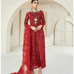 May include: A red formal dress with long sleeves and intricate embroidery. The dress features a high neckline and a slit up the front. A matching sheer scarf with fringe detail drapes from the shoulder. The model is wearing jewelry and nude heels.