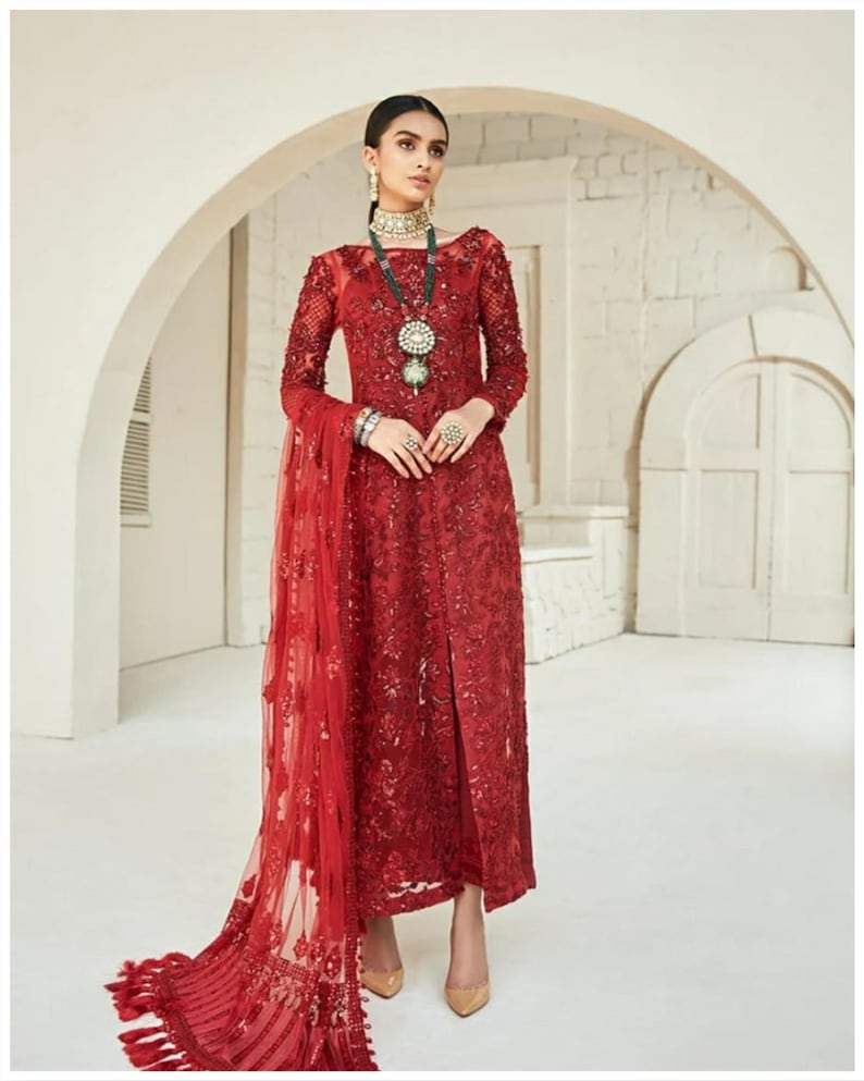May include: A red formal dress with long sleeves and intricate embroidery. The dress features a high neckline and a slit up the front. A matching sheer scarf with fringe detail drapes from the shoulder. The model is wearing jewelry and nude heels.