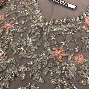 以下が含まれることがあります： 複雑な刺繍が施されたグレーの生地のクローズアップ。デザインは、銀と金の糸、スパンコール、小さな真珠のようなビーズが特徴です。ピンクの花柄のアクセントがパターン全体に散りばめられています。「NECK」という単語が白いタグに印刷されています。