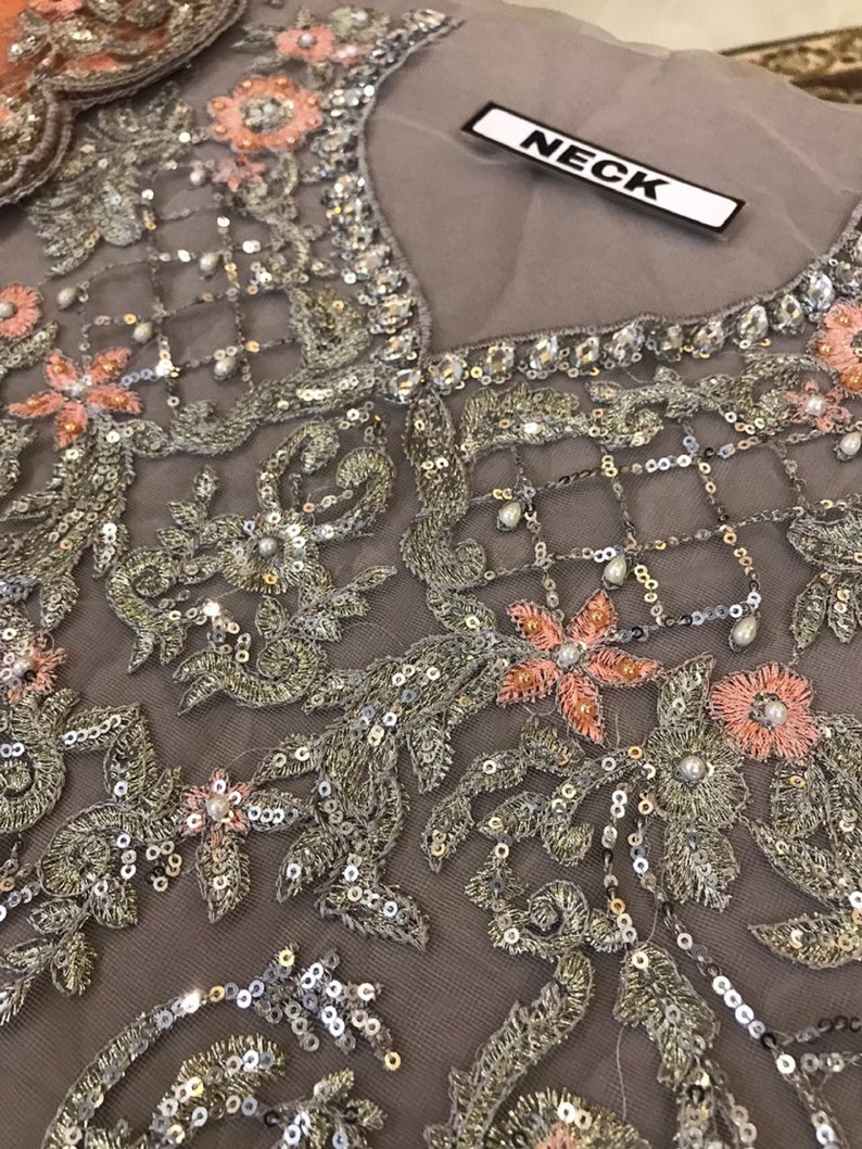 以下が含まれることがあります： 複雑な刺繍が施されたグレーの生地のクローズアップ。デザインは、銀と金の糸、スパンコール、小さな真珠のようなビーズが特徴です。ピンクの花柄のアクセントがパターン全体に散りばめられています。「NECK」という単語が白いタグに印刷されています。