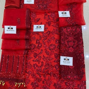 May include: A red embroidered fabric set, including a trouser, dupatta, and various panels labeled "front," "back," and "sleeves." The fabric features intricate floral patterns and embellishments.