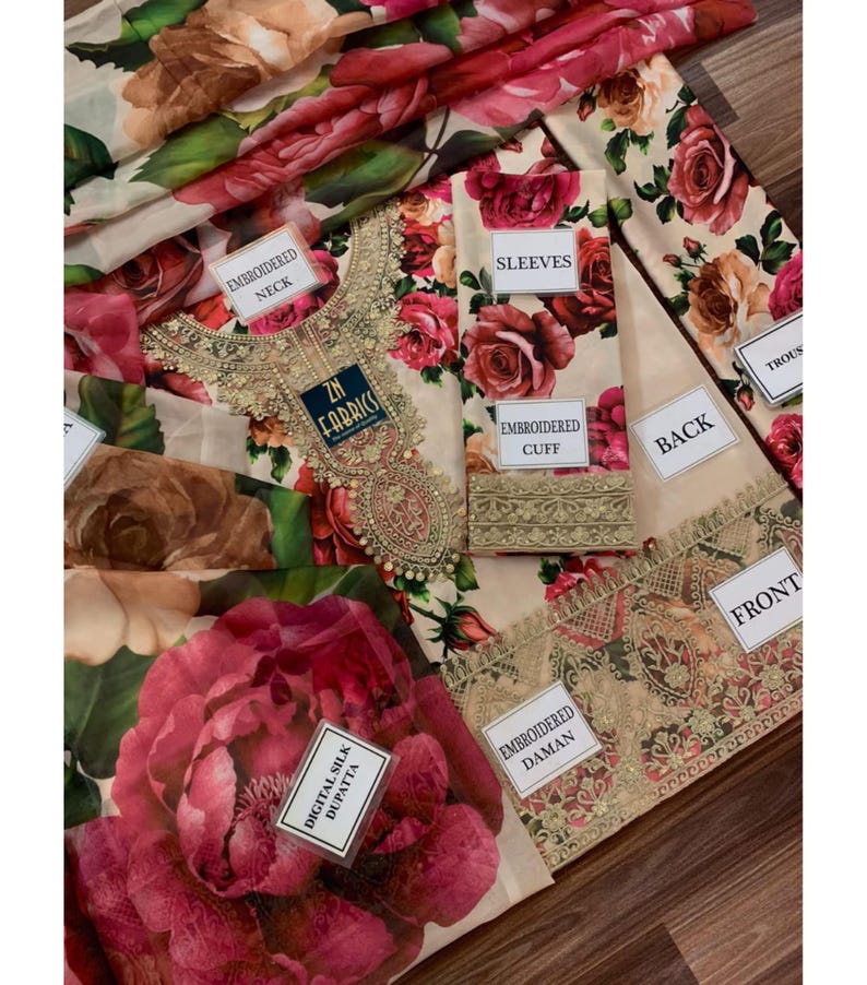 Puede incluir: Un conjunto de tela con estampados florales y etiquetas que indican "Embroidered Neck", "Sleeves", "Embroidered Cuff", "Back", "Front" y "Embroidered Daman". El dise&ntilde;o presenta grandes rosas rojas y rosas sobre un fondo crema.