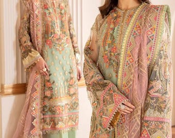 May include: Two women wearing intricately embroidered dresses. The dresses are a light green color with floral and geometric patterns in pink, gold, and brown. The dresses have long sleeves and are paired with matching scarves. The outfits are elegant and formal.