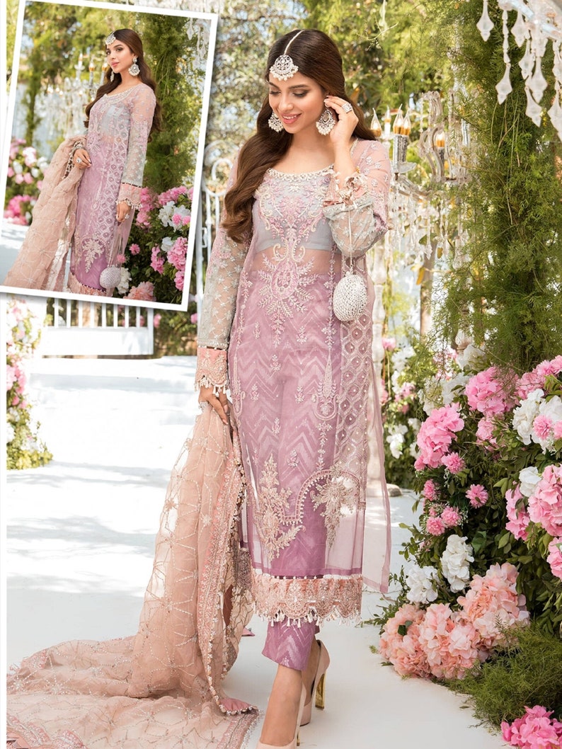 May include: A sheer, light purple formal dress with intricate embroidery and long sleeves. The dress features a matching scarf and is paired with light pink heels. The background includes pink and white flowers and decorative lighting.