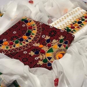 Könnte beinhalten: Zweiteiliges besticktes Stoffset. Ein Teil ist tiefrot mit einem geometrischen Muster und bunten Pompon-Akzenten. Das andere Teil ist weiß mit einem Zickzackmuster und bunten Akzenten. Weißer Stoff drapiert die Teile.