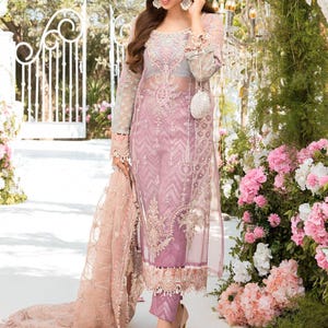 May include: A sheer, light purple embroidered dress with long sleeves and matching pants. The dress features intricate floral patterns and is paired with high-heeled shoes. A matching scarf is draped over the arm. The background includes a white gate and pink flowers.