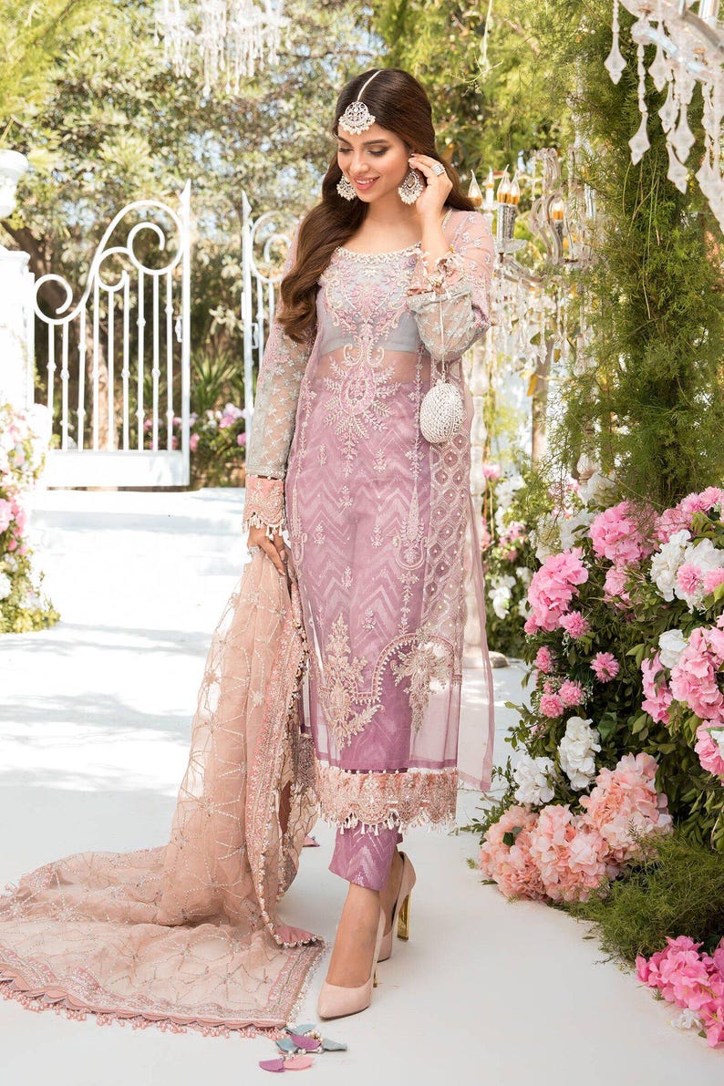 May include: A sheer, light purple embroidered dress with long sleeves and matching pants. The dress features intricate floral patterns and is paired with high-heeled shoes. A matching scarf is draped over the arm. The background includes a white gate and pink flowers.