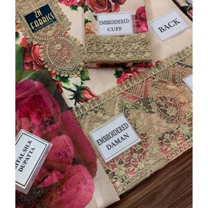 Puede incluir: Una tela beige con un estampado floral de rosas rojas y rosas. La tela tiene detalles bordados y etiquetas que dicen "EMBROIDERED CUFF", "BACK", "ITAL SILK DUPATTA" y "EMBROIDERED DAMAN". La tela es de "ZN FABRICS".