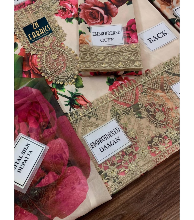 Puede incluir: Una tela beige con un estampado floral de rosas rojas y rosas. La tela tiene detalles bordados y etiquetas que dicen "EMBROIDERED CUFF", "BACK", "ITAL SILK DUPATTA" y "EMBROIDERED DAMAN". La tela es de "ZN FABRICS".