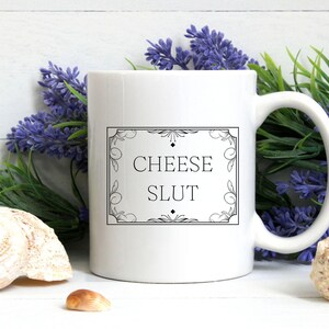 May include: A white ceramic mug featuring a black rectangular border with decorative corner flourishes and the text "CHEESE SLUT" printed in the centre. The mug has a standard handle and a smooth, glossy finish on its exterior surface.