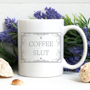 Pu&ograve; includere: Tazza da caff&egrave; in ceramica bianca con un contorno nero di una cornice decorativa e il testo "COFFEE SLUT" all'interno della cornice.