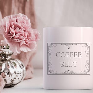 Pu&ograve; includere: Tazza da caff&egrave; in ceramica bianca con un contorno nero di una cornice decorativa e il testo "COFFEE SLUT" all'interno della cornice.