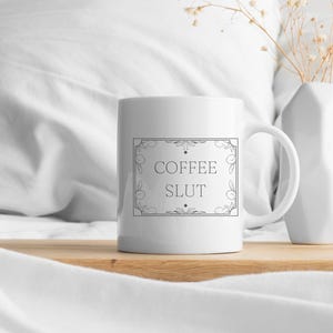 Pu&ograve; includere: Tazza da caff&egrave; in ceramica bianca con un contorno nero di una cornice decorativa e il testo "COFFEE SLUT" all'interno della cornice.