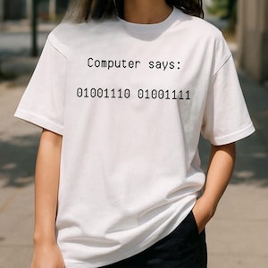 May include: White t-shirt with the text "Computer says:" above a line of binary code. The shirt has short sleeves and a crew neck. The person is wearing black trousers.