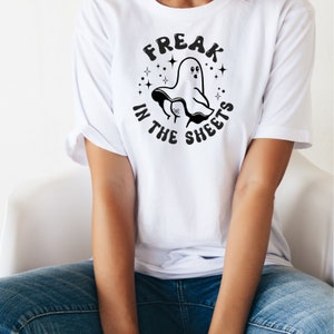 May include: White t-shirt featuring a black graphic of a ghost and the text "FREAK IN THE SHEETS" in a circular design. The ghost has a surprised expression and is surrounded by stars. The shirt is worn by a person.