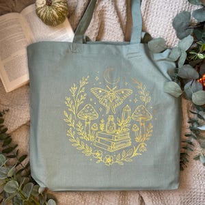 May include: A sage green tote bag featuring a gold-colored line art illustration of a moth, mushrooms, a stack of books, a small bottle, and a crescent moon, all surrounded by a circular arrangement of leafy branches and small flowers.