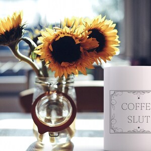Pu&ograve; includere: Una tazza da caff&egrave; in ceramica bianca con un contorno nero di una cornice decorativa e il testo "COFFEE SLUT" all'interno. La tazza &egrave; appoggiata su una superficie bianca con un mazzo di girasoli in un barattolo di vetro.