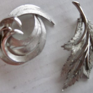 May include: Two silver tone brooches. One is a swirl design and the other is a leaf design.
