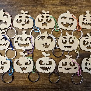 May include: A set of 14 wooden pumpkin keyrings with different facial expressions. Each keyring is painted in a natural wood colour and has a metal ring attached.