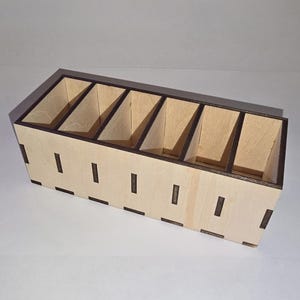 Peut inclure: Un organisateur en bois de couleur claire avec six compartiments. L'organisateur a une forme rectangulaire avec des bordures marron foncé et des détails découpés sur les côtés. Les compartiments sont inclinés.
