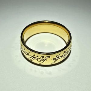 以下が含まれることがあります： 黒い刻印がバンドの周りに施された、金色のリング。リングは円形で、金属製に見えます。スクリプトは様式化されたフォントです。リングは白い表面にあります。