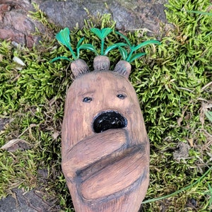 May include: A brown ceramic sculpture of a creature with a surprised expression. The creature has a long, thin body with a rounded head and a small mouth. It has a few green leaves growing from the top of its head. The sculpture is sitting on a bed of green moss.