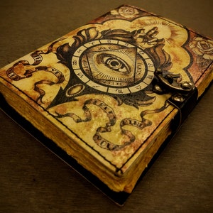 May include: A vintage-style leather-bound journal with an all-seeing eye design. The cover features an eye inside a triangle, surrounded by a circular compass with Roman numerals and zodiac symbols. The journal has a leather strap closure and a decorative metal clasp.
